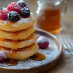 A stack of golden mini pancakes topped with syrup and fresh berries on a rustic table.